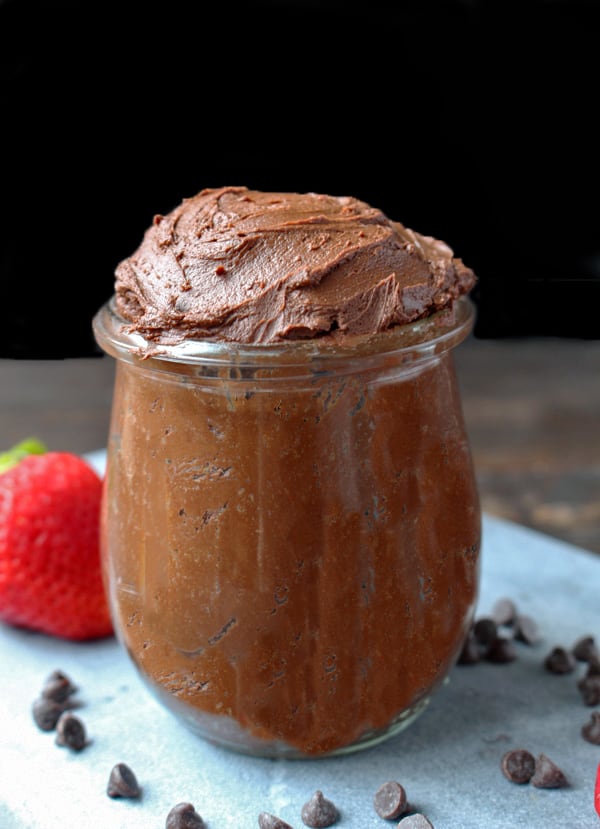Paleo chocolate frosting in a clear jar with a strawberry in the background. 