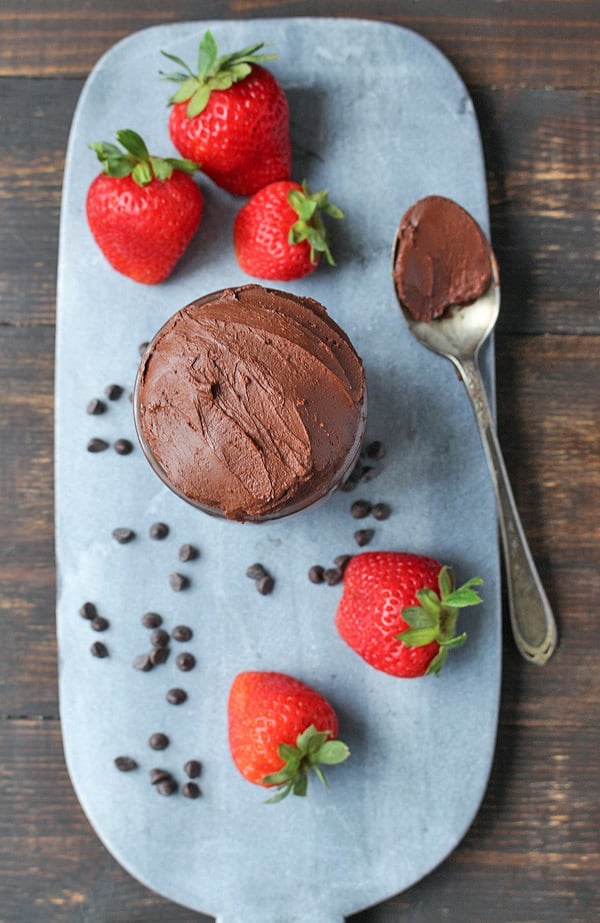 paleo chocolate frosting on a stone cutting board with strawberries and chocolate chips surrounding it. 
